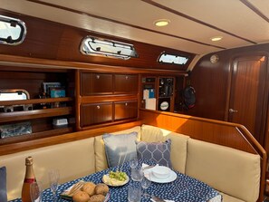A boat cabin with a dining area featuring a table set for two, a cushioned bench, and a wooden cabinet with various items.