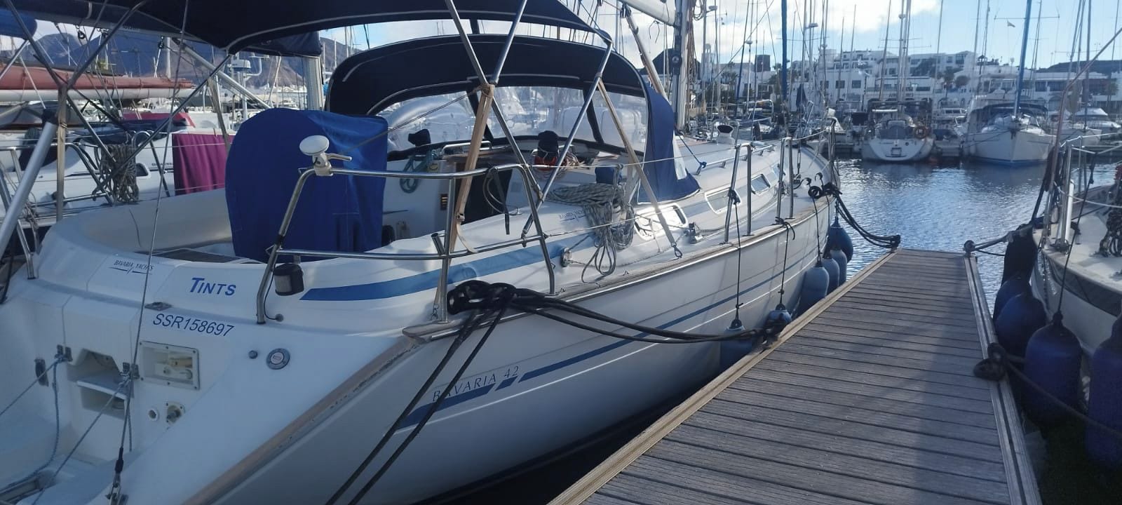 En segelbåt låg förtöjd vid en småbåtshamn, med ett trädäck och andra båtar i bakgrunden.