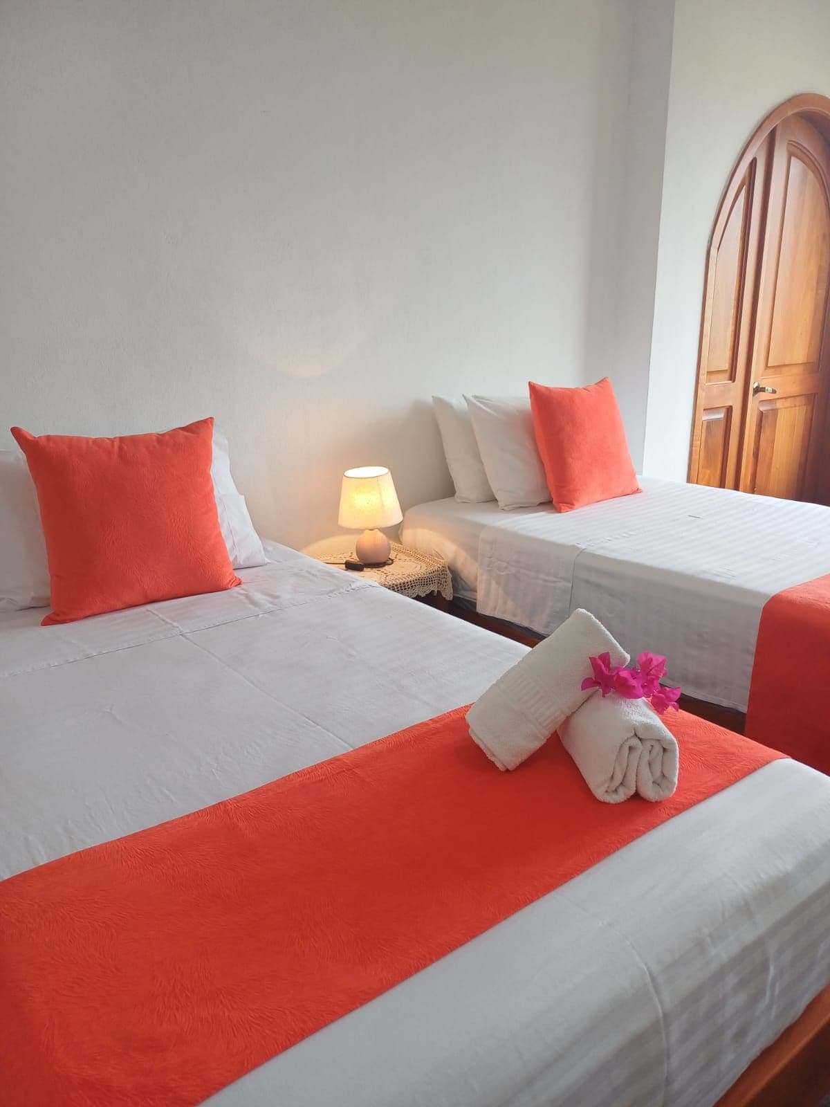Habitación de hotel con dos camas, cada una con un cojín naranja decorativo, una lámpara de noche y una toalla doblada con flores sobre la cama.