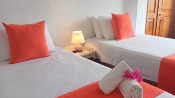 Habitación de hotel con dos camas, cada una con un cojín naranja decorativo, una lámpara de noche y una toalla doblada con flores sobre la cama.