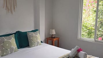 Una cama bien hecha con una colcha estampada, dos almohadas con diseños circulares, un tapiz decorativo y una ventana con vista a follaje.