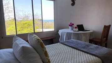 Habitación de hotel con cama, escritorio con una computadora portátil, silla, ventana con vista a árboles y al mar, y un adorno de pared.