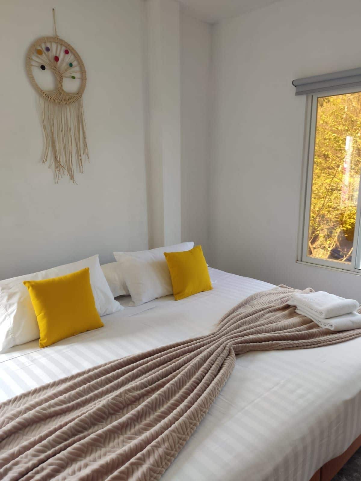 Una cama con ropa de cama blanca, dos almohadas amarillas y un plaid color beige, junto con un tapiz con diseño de árbol.