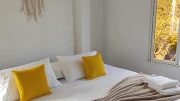 Una cama con ropa de cama blanca, dos almohadas amarillas y un plaid color beige, junto con un tapiz con diseño de árbol.