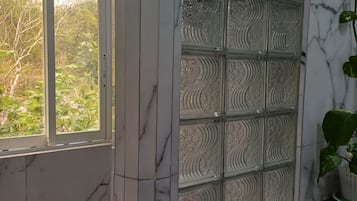 A bathroom with a glass partition, marble walls, and a window with a view of trees.