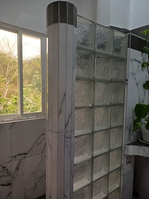 A bathroom with a glass partition, marble walls, and a window with a view of trees. - Bahía Garden (Puerto Ayora)