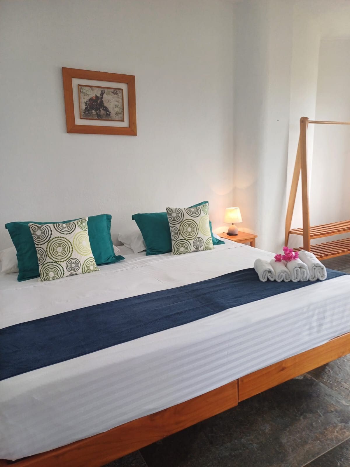 Cama bem arrumada com colcha azul e almofadas verdes, cabeceira de madeira, mesa de cabeceira com abajur e quadro na parede.