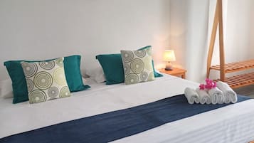Cama bem arrumada com colcha azul e almofadas verdes, cabeceira de madeira, mesa de cabeceira com abajur e quadro na parede.