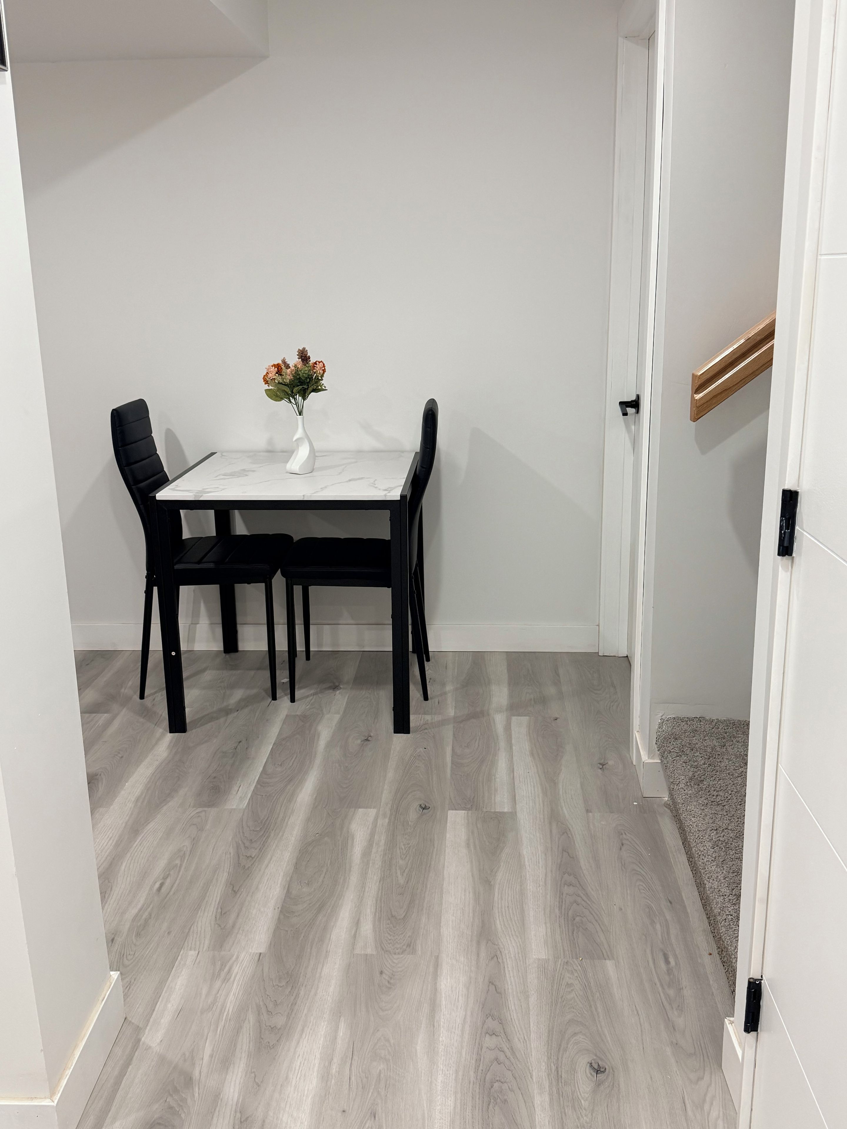 Een eetgedeelte met een witte tafel en zwarte stoelen, een vaas met bloemen, en een houten vloer.