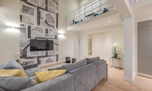 Een ruime woonkamer met een grijze bank, een flatscreen televisie aan de muur en een trap naar een hoger gelegen verdieping.