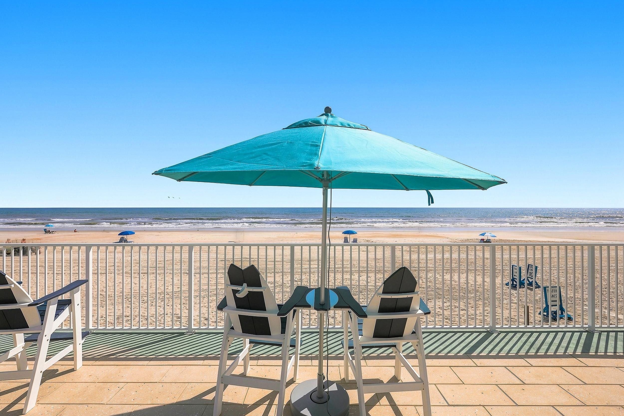 A beachside seating area with a turquoise umbrella, chairs, and a view of the ocean.