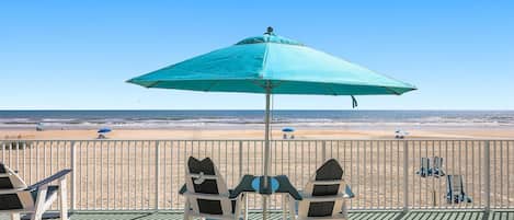 A beachside seating area with a turquoise umbrella, chairs, and a view of the ocean.