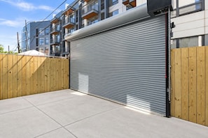 A closed garage door with a metal roller shutter, a wooden fence, and a modern building with balconies in the background. - Modern 4BR/3 bath | Rooftop Deck, Parking &Sleeps 12 (Washington)
