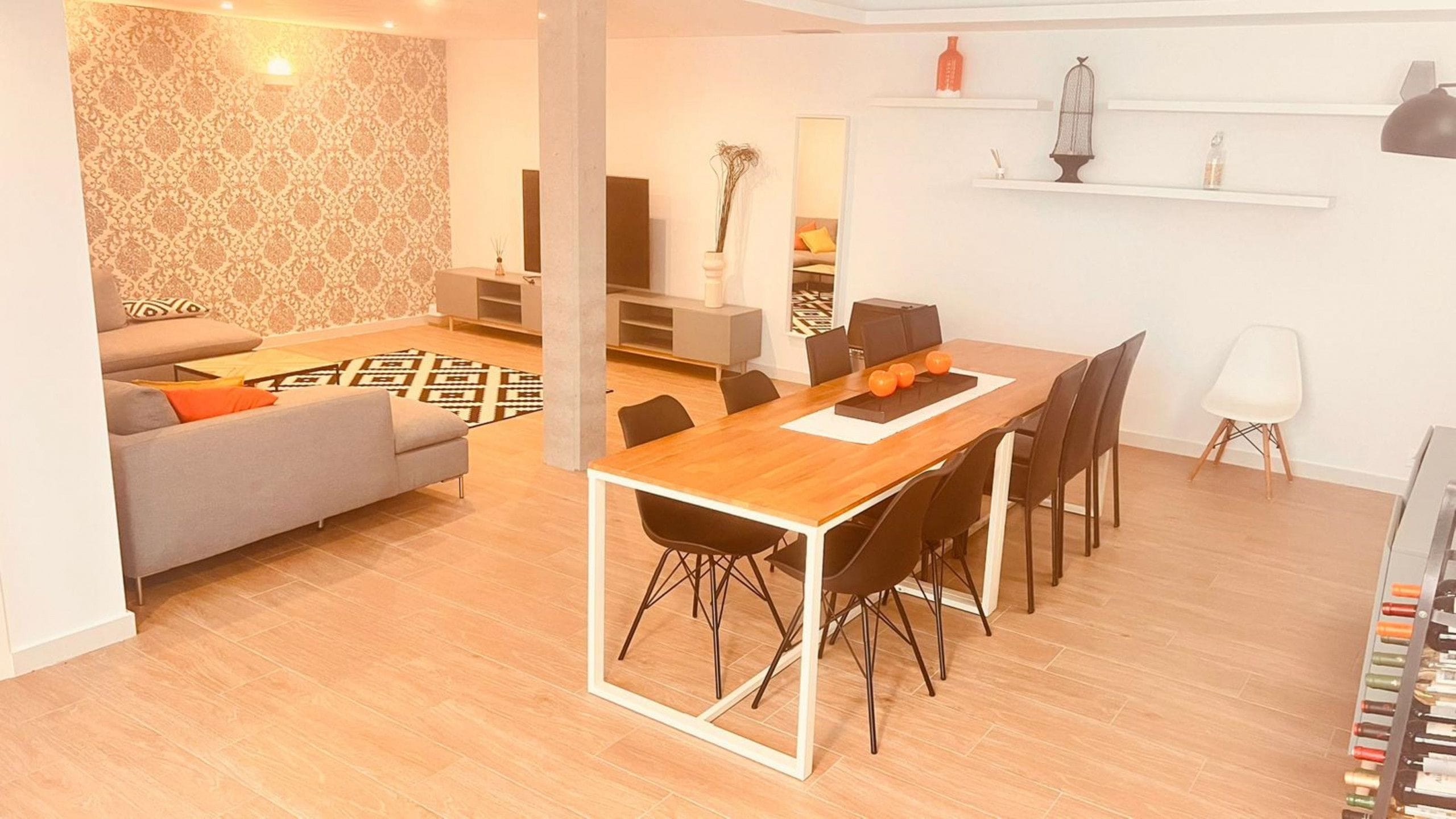 A modern living room with a sofa, coffee table, and patterned wallpaper. A dining area with a wooden table and chairs is visible.