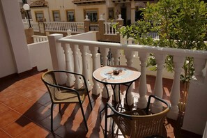 Ein Balkon mit zwei Korbstühlen, einem kleinen runden Tisch, weißen Brüstungen und einem Blumentopf.