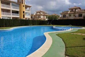 A swimming pool with a curved edge, surrounded by a green lawn and a hedge. - Apartment La Zenia Rubi (Orihuela)