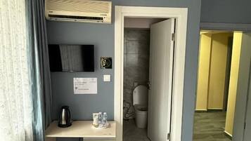 A hotel room with a desk, a bed, an air conditioning unit, a television, and a bathroom visible through an open door.