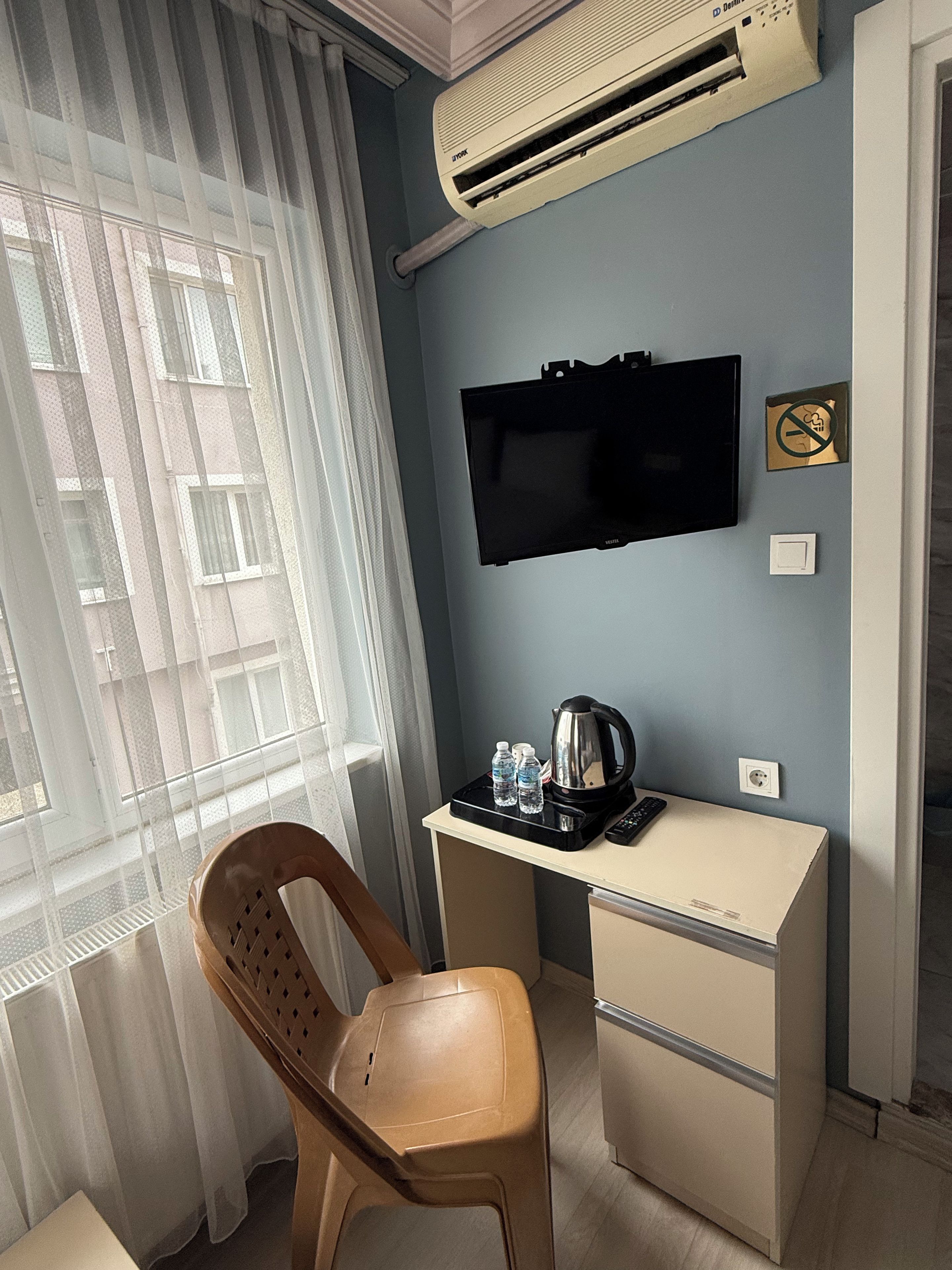 A hotel room with a desk, chair, television, air conditioning unit, and a view of the outside.