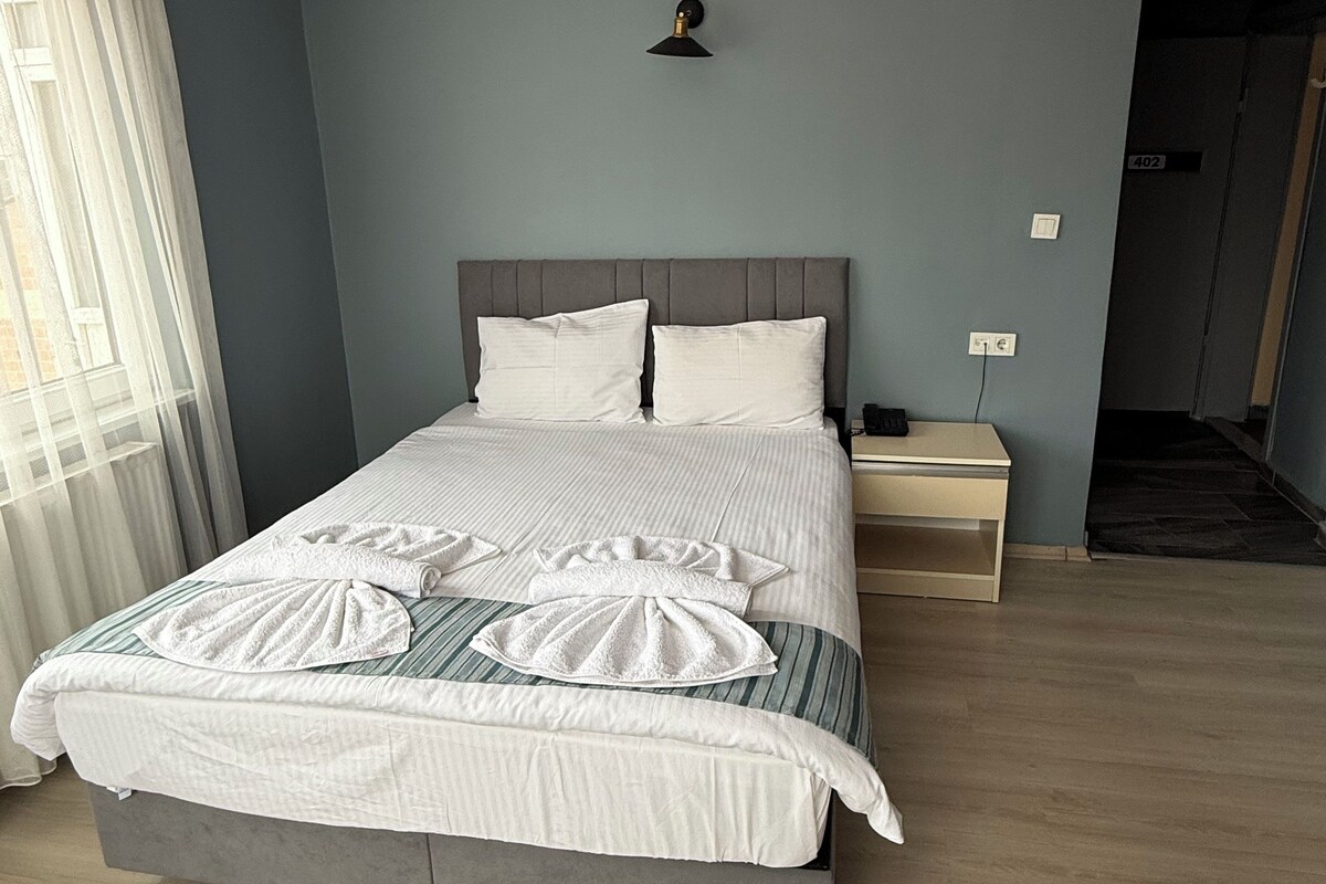 A neatly made bed with white linens and a towel arranged on the bed. There is a bedside table with a lamp and a power outlet. The room has light blue walls and a wooden floor.