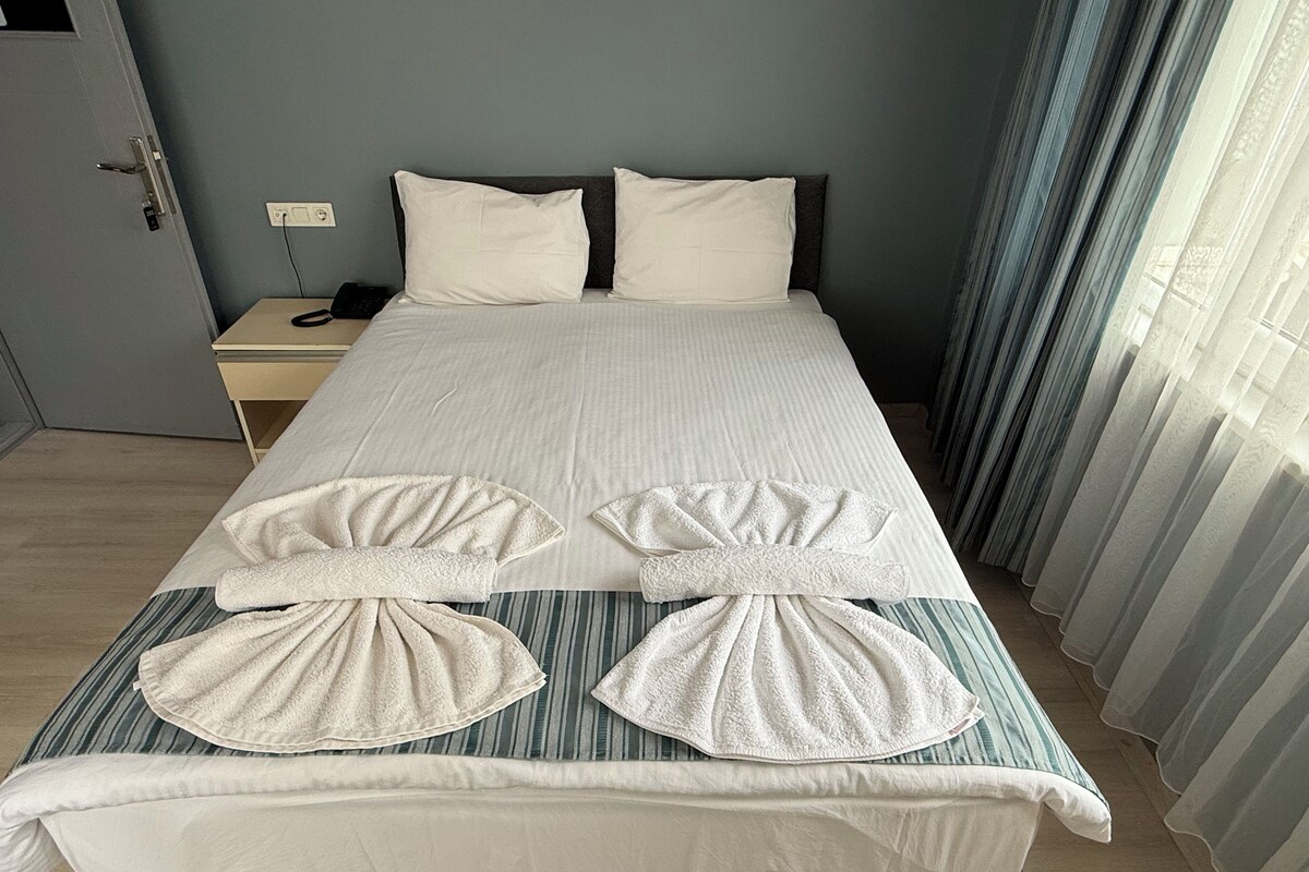 A hotel room with a bed, pillows, a bedside table with a phone, and a window with curtains.