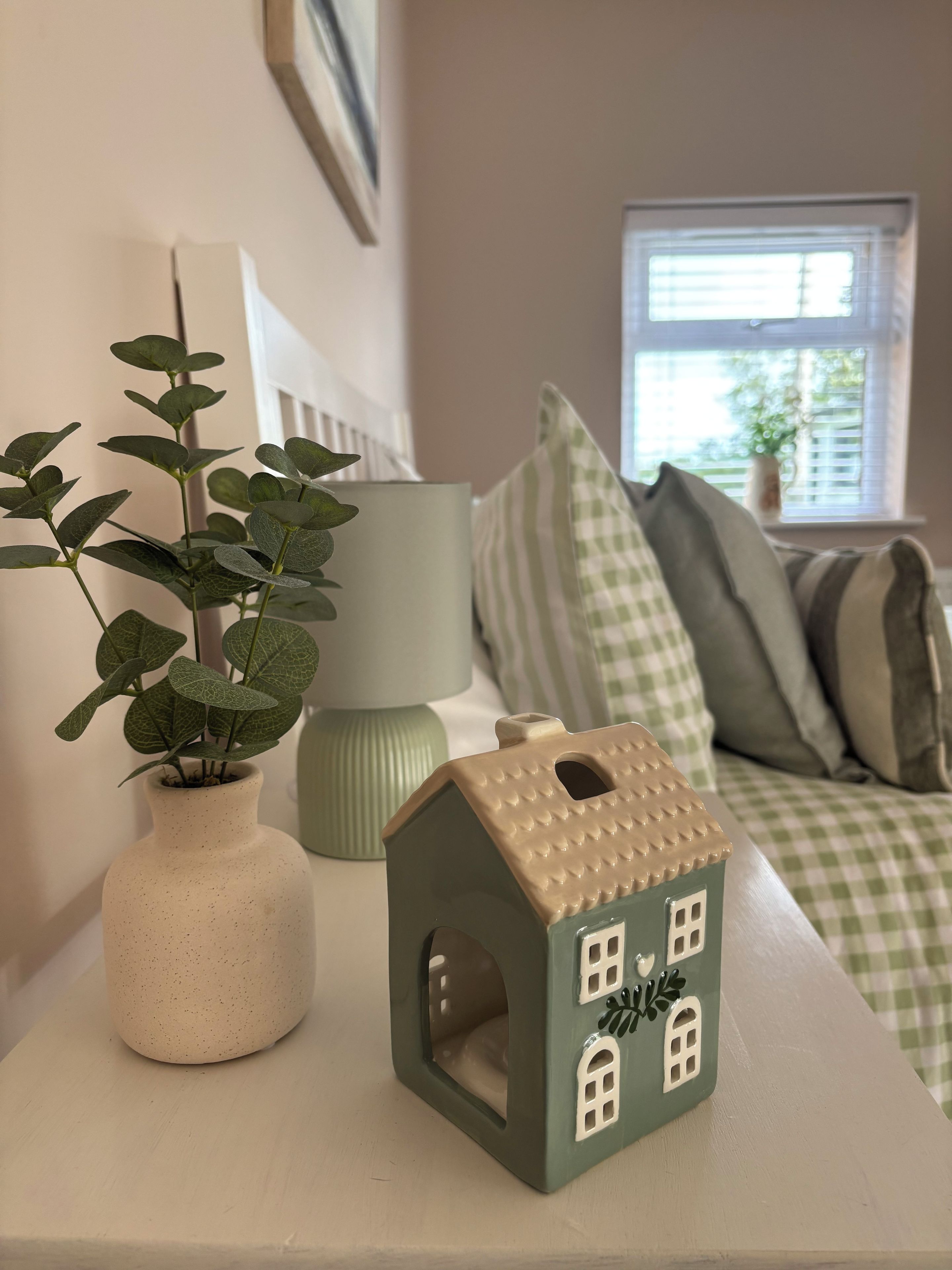 A small house-shaped object on a surface, a plant in a vase, and a window with blinds in the background.