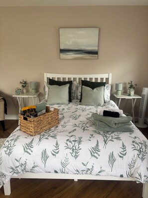 A neatly made bed with a floral-patterned bedspread, two bedside tables with lamps, a small house-shaped decoration, and a wall-mounted painting.