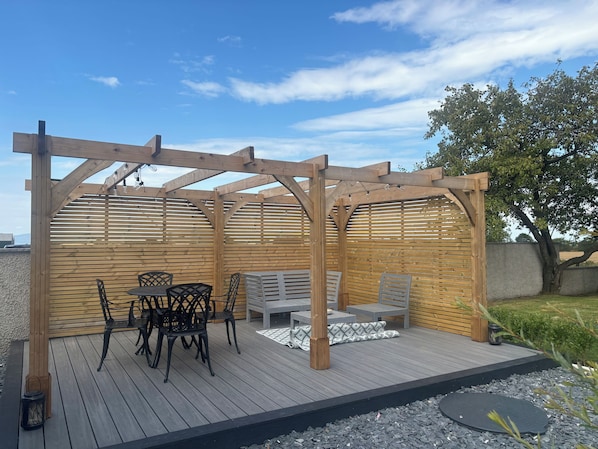 A wooden pergola with a table and chairs, surrounded by a gravel area and a wooden deck.