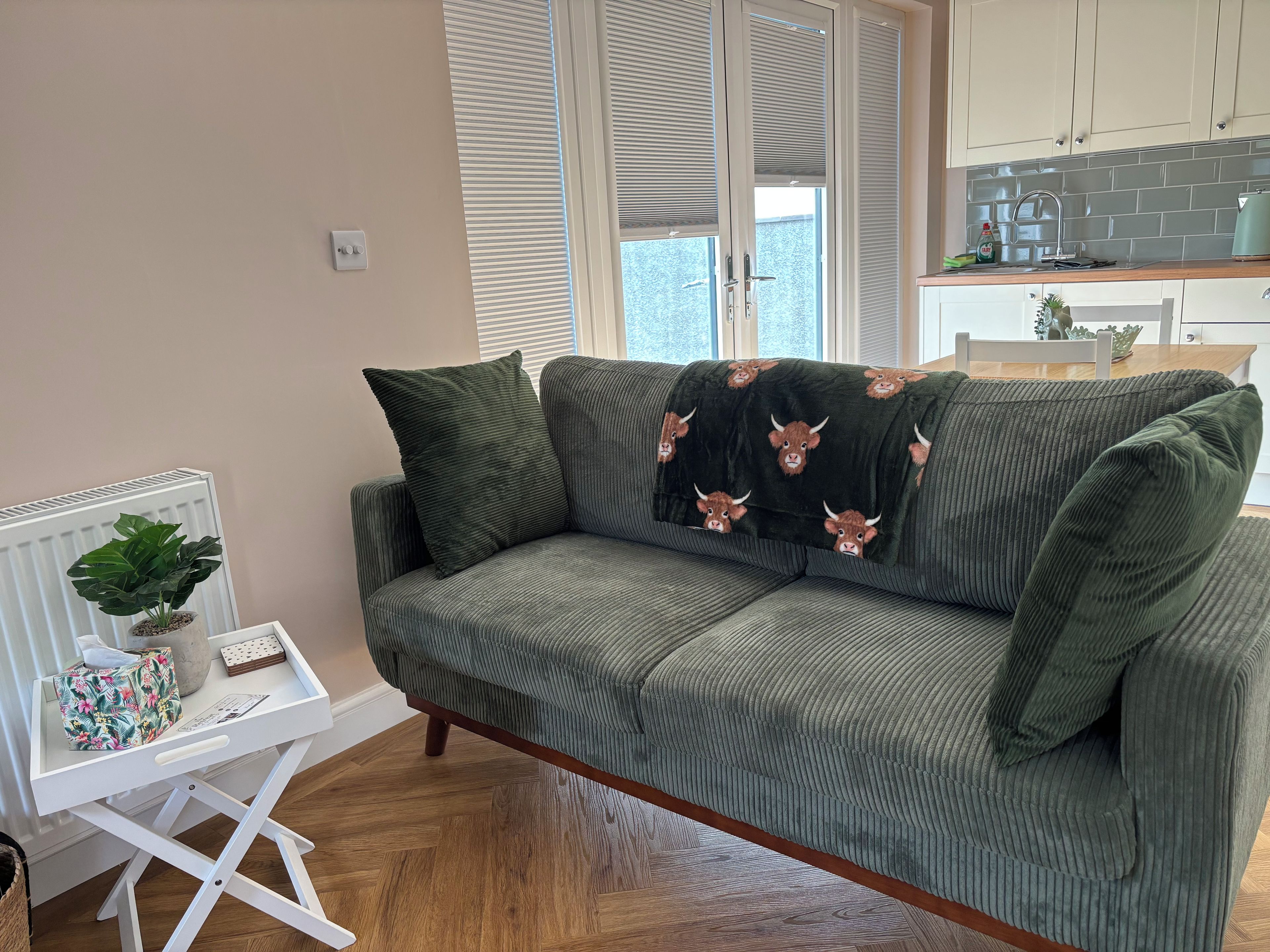 A green sofa with a bull-themed throw, a white side table with a plant and a floral bag, and a kitchen area with a tiled backsplash and cabinets.