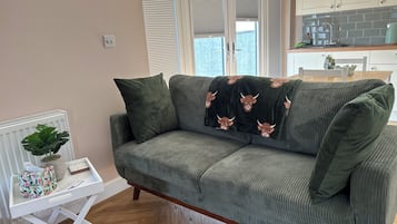 A green sofa with a bull-themed throw, a white side table with a plant and a floral bag, and a kitchen area with a tiled backsplash and cabinets.