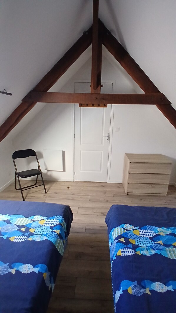 A small, cozy attic bedroom with two beds, a chair, a dresser, and a door. - Vacation Rental at Lake Guerlédan (Saint-Aignan)