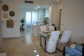 A dining area with a glass table and chairs, a kitchen in the background, and a potted plant. - Secreto La Zenia (Orihuela)