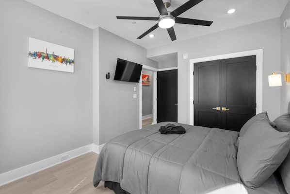 Un dormitorio con cama, ventilador de techo, televisor de pantalla plana, una puerta y una lámpara de pared.