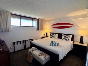 Quarto de hotel com cama, luminárias de cabeceira, janela com cortinas, um quadro na parede e uma pequena mesa com abajur.