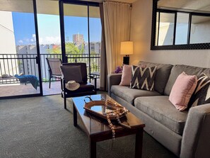 A living room with a sofa, a coffee table, and a chair. There is a balcony with a view of buildings and palm trees. - Ocean View Kaanapali Shores Spacious One Bedroom (Lahaina)