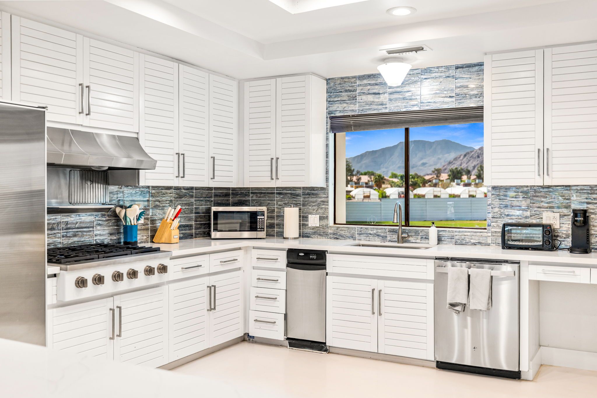 A modern kitchen with white cabinetry, stainless appliances, and a view of mountains through a large window.