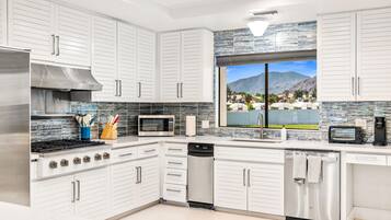 A modern kitchen with white cabinetry, stainless appliances, and a view of mountains through a large window.
