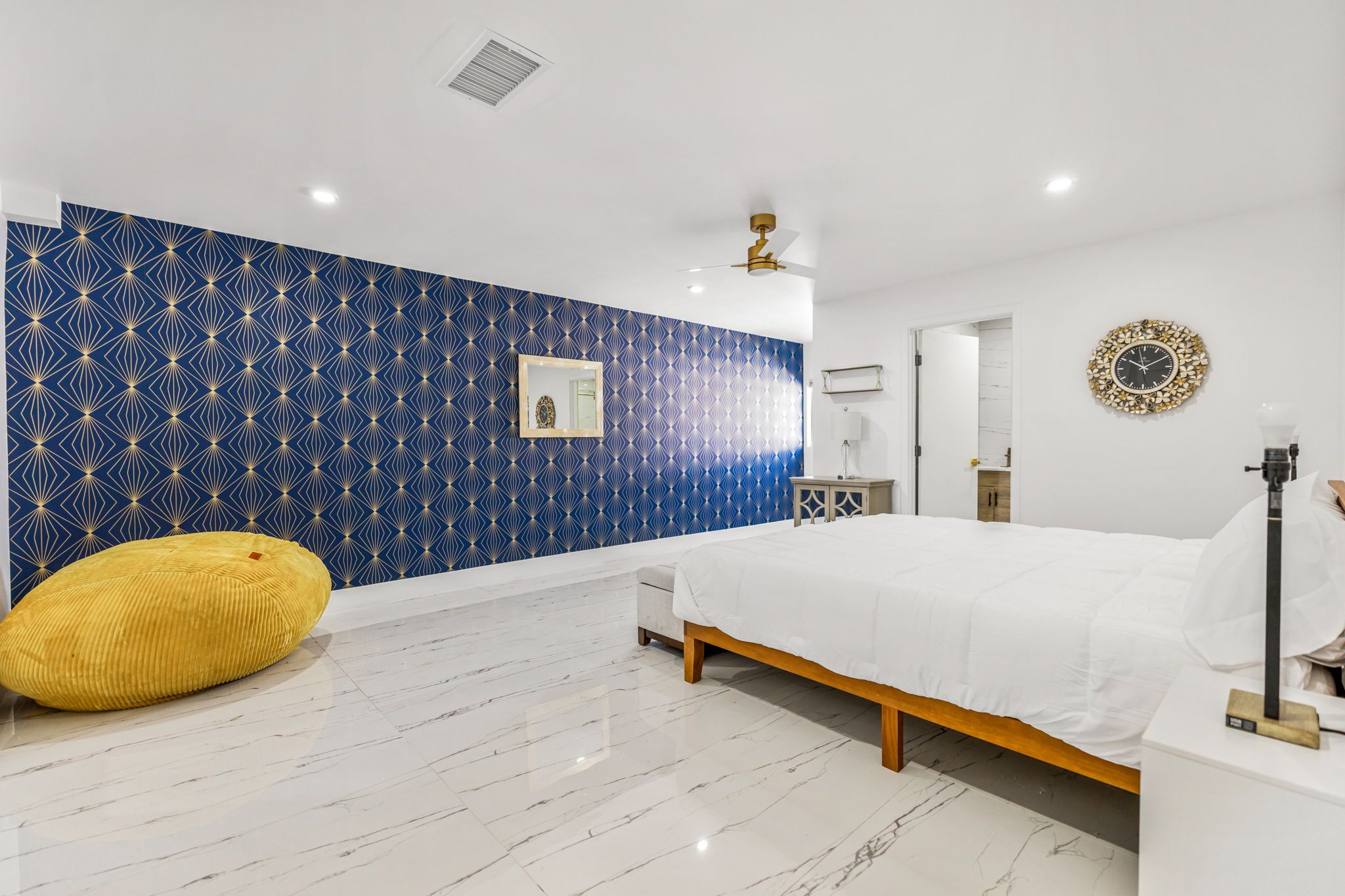 A modern bedroom with a patterned blue wall, a white bed, a yellow bean bag, and a ceiling fan.