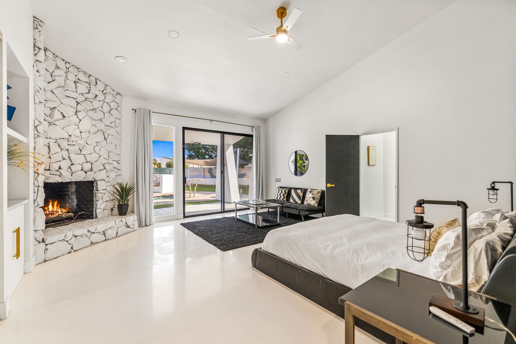 A modern bedroom with a stone fireplace, a bed, a desk, and a view to an outdoor patio.