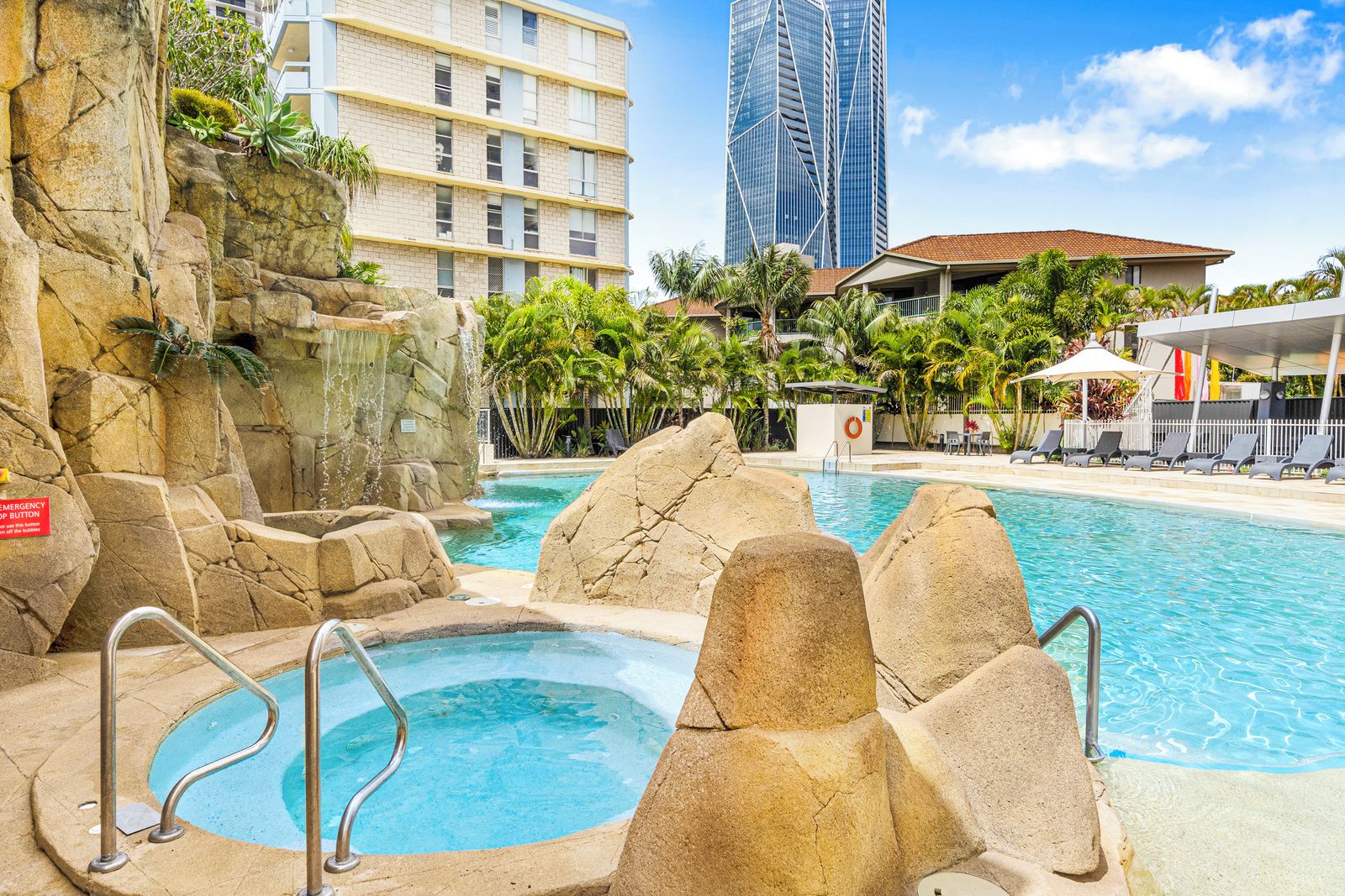 A hotel pool area with a hot tub, a rock wall, and a view of a tall building.