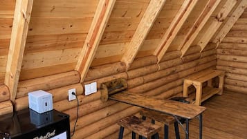 Interior de una cabaña de troncos con piso de madera, un mueble negro con un cartel, una pequeña mesa de madera y un banco.