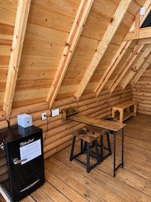 A log cabin interior with a wooden floor, a black cabinet with a sign, a small wooden table, and a bench.