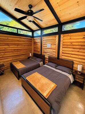 A log cabin bedroom with two beds, a ceiling fan, and a window with a view of trees. - VILLA AMADA. Cabaña Agave (Santiago)