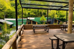 An outdoor patio area with wooden tables and benches, surrounded by a wooden railing, and overlooking a lush green landscape.