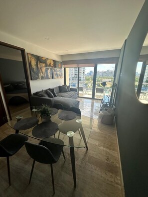 Un salon moderne avec une table en verre, des fauteuils foncés, un canapé gris et une vue sur la ville à travers de grandes fenêtres.