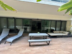 Une terrasse avec une table et deux fauteuils, un canapé et une porte-fenêtre coulissante donnant sur un espace intérieur.