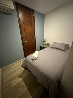 A bedroom with a bed, a wooden door, a nightstand with a lamp, and a wall-mounted air conditioner. - El Encuentro. Tu mejor opción para viajar. (Cancún)
