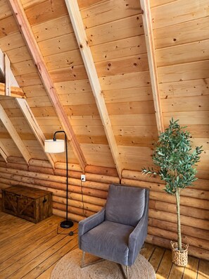 A log cabin interior with a sloped ceiling, a gray upholstered chair, a floor lamp, and a potted plant.