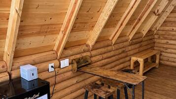 A log cabin interior with a wooden ceiling, a small table, a bench, and a black refrigerator.