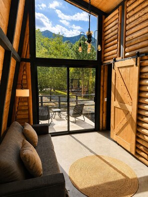 Interior d'una cabana de fusta moderna amb un sofà, un otomanet trenat i vistes a un pati amb mobles d'exterior.