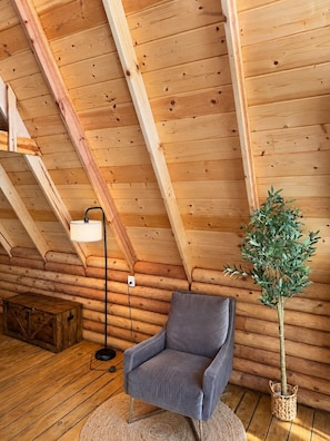 Interior de una cabaña de troncos con techo inclinado, un sillón tapizado gris, una lámpara de pie y una planta en maceta.